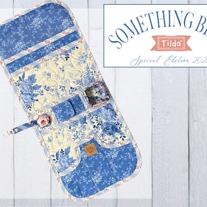 May include: A quilted fabric organizer featuring a blue and cream floral pattern with multiple pockets, a small square container, and a strap with a floral button. The text SOMETHING BLUE Tilda Special Edition 2026 is displayed in a rectangular box.