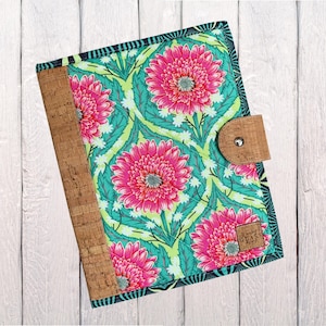 May include: A floral journal with a cork spine and closure. The cover features a pattern of large pink gerbera daisies on a teal background with green leaves. The journal has a cork closure and spine, and a small label that reads "Handmade Glass".