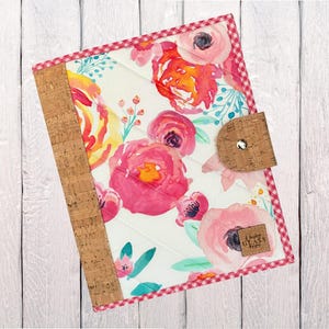 May include: A floral journal with a cork spine and closure. The cover features a quilted white background with pink, orange, and yellow watercolor flowers. The edges are trimmed with a red and white gingham pattern.