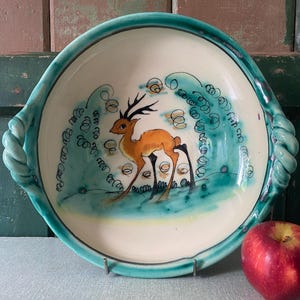 May include: A ceramic serving bowl with a deer illustration. The bowl has a white interior with a teal rim and handles. The deer is orange with black antlers, standing in a teal and white landscape. A red apple rests on the table.