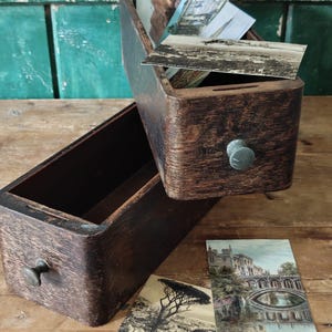 May include: A dark brown wooden card catalogue drawer with a small metal knob, containing a collection of vintage postcards. Two additional postcards are placed outside the drawer, one featuring a tree and the other a stone bridge over water.