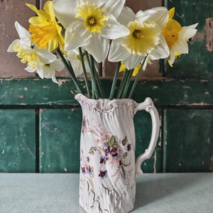 Peut inclure: Un pichet en céramique blanche orné de motifs floraux contient un bouquet de jonquilles. Les jonquilles ont des pétales blancs et des centres jaunes. Le pichet est décoré de fleurs roses, violettes et vertes.