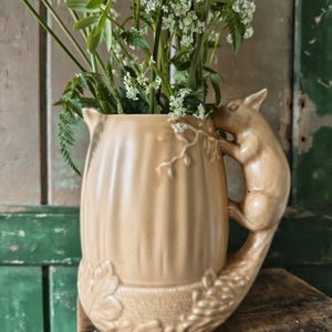May include: A tan ceramic jug with a squirrel handle and leaf detailing. The jug is filled with white wildflowers and green foliage, creating a rustic display. The jug is approximately 20 cm tall.