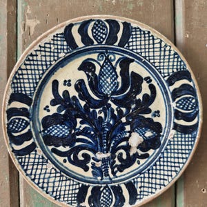 May include: A round, antique ceramic plate with a white base and intricate blue floral and geometric patterns. The plate's rim features a crosshatch design, and the central motif is a stylized flower. The plate has a rustic, aged appearance.