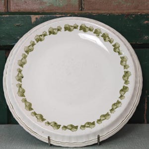 May include: A round, off-white ceramic plate featuring a decorative border of small, repeating green fish illustrations. The plate has a slightly scalloped, raised edge and a smooth, glossy finish, presenting a clean and simple design.