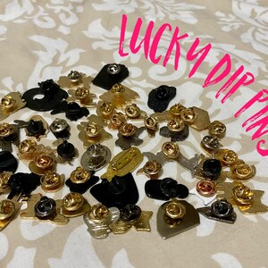 May include: A collection of colorful enamel pins with various designs, including hearts, stars, and abstract shapes. The pins are arranged on a beige fabric background. The text "LUCKY DIP PINS" is written in pink letters on the image.