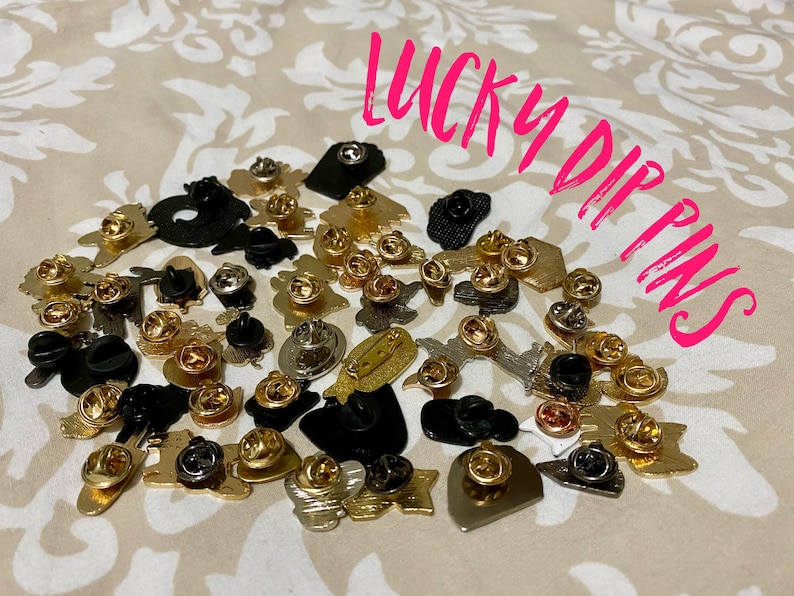 May include: A collection of colorful enamel pins with various designs, including hearts, stars, and abstract shapes. The pins are arranged on a beige fabric background. The text "LUCKY DIP PINS" is written in pink letters on the image.