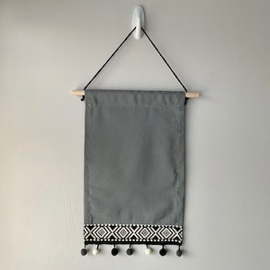 May include: Grey wall tapestry with black and white geometric pattern and white pom pom trim.