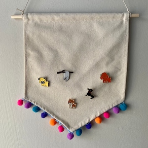 May include: A beige canvas banner with a pom-pom trim hangs from a wooden dowel. Five enamel pins featuring different dog breeds are pinned to the banner.