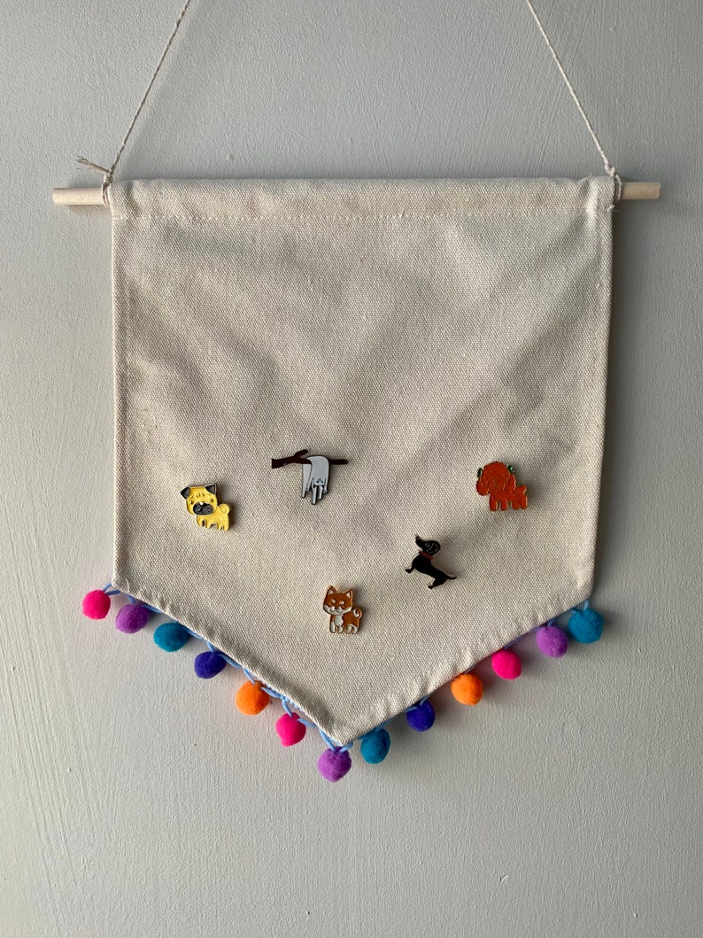 May include: A beige canvas banner with a pom-pom trim hangs from a wooden dowel. Five enamel pins featuring different dog breeds are pinned to the banner.