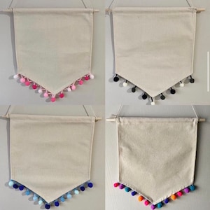 May include: Four blank canvas pennants with different colored pom pom trim. The pennants are hanging from a wooden dowel rod. The pom pom colors are pink, black and white, blue, and orange and yellow.