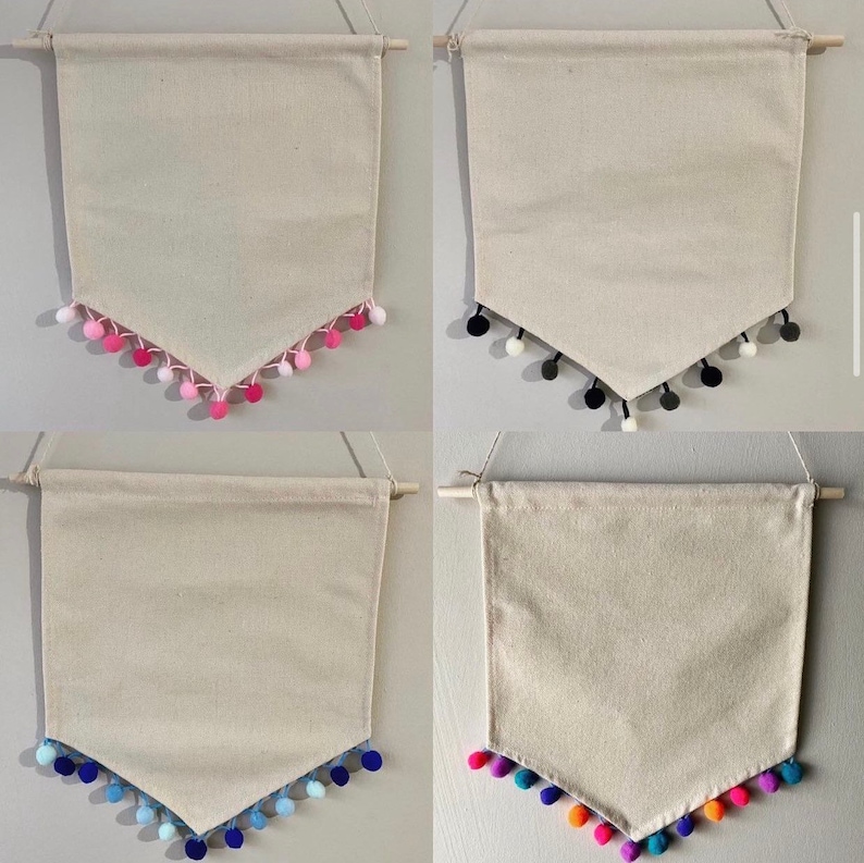 May include: Four blank canvas pennants with different colored pom pom trim. The pennants are hanging from a wooden dowel rod. The pom pom colors are pink, black and white, blue, and orange and yellow.