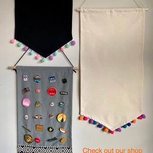 May include: Three fabric wall banners with pom-pom trim. The top banner is black, the middle banner is gray with a variety of pins and badges, and the bottom banner is white. The text on the bottom banner reads "Check out our shop for more styles, colours and sizes!"