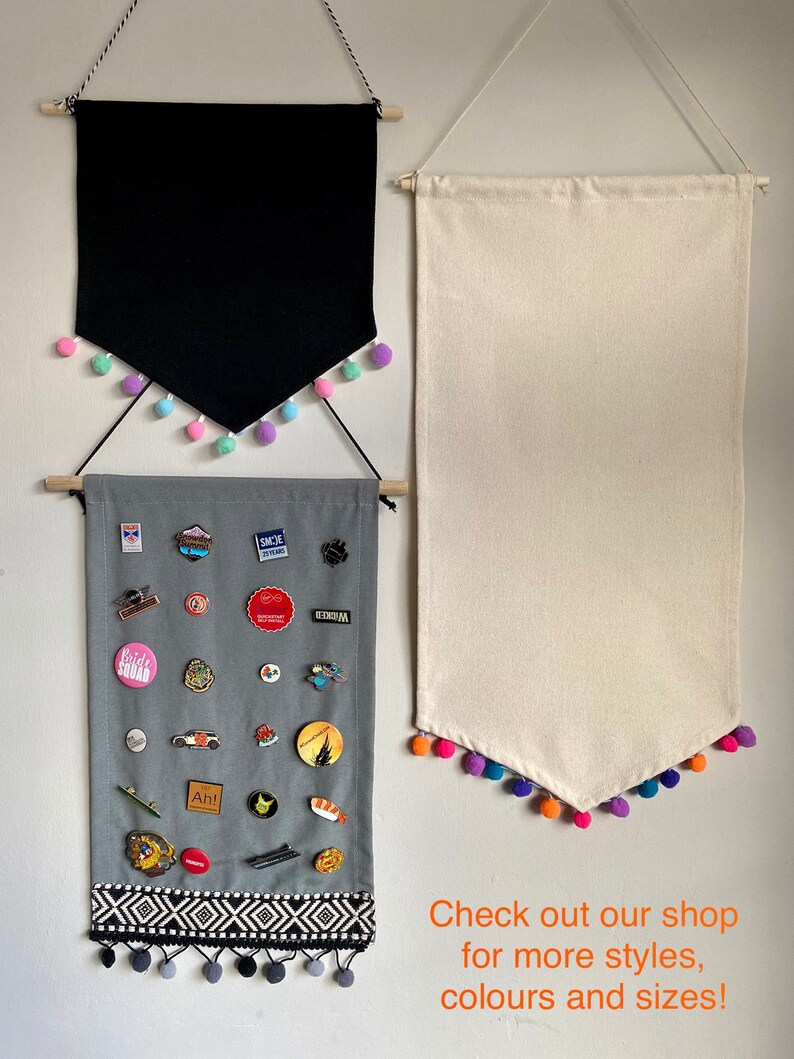 May include: Three fabric wall banners with pom-pom trim. The top banner is black, the middle banner is gray with a variety of pins and badges, and the bottom banner is white. The text on the bottom banner reads "Check out our shop for more styles, colours and sizes!"