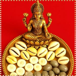 May include: A gold-coloured statue of a seated deity holding lotus flowers, on a gold tray with seashells, spiral shells, and grey stones. The background is a solid red colour with a white and silver border.