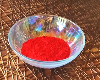 May include: A clear, iridescent glass bowl filled with a vibrant red powder. The bowl sits on a brown, textured surface. The powder appears finely ground and evenly distributed within the bowl.