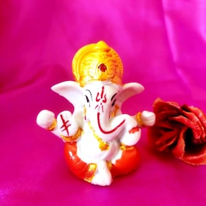 Könnte beinhalten: Kleine, weiß-orangefarbene Figur des hinduistischen Gottes Ganesha, sitzend mit ausgestreckten Armen. Die Figur hat eine goldene Krone und rote Akzente. Rechts eine rote Blume, vor pinkfarbenem Hintergrund.