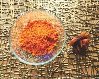 May include: A small glass bowl filled with bright orange powder, likely a spice like paprika or turmeric. The bowl is sitting on a gold-colored woven surface.