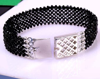 May include: A black beaded bracelet with a silver clasp. The bracelet is made of small, round beads that are woven together in a pattern. The clasp is a simple, rectangular design with a decorative pattern.