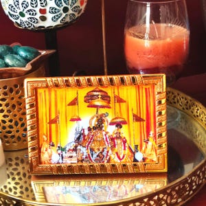 May include: A framed religious image with a gold border. The image depicts figures in ornate clothing, set against a yellow backdrop with red accents. The frame sits on a gold-colored tray, with a candle and decorative items in the background.
