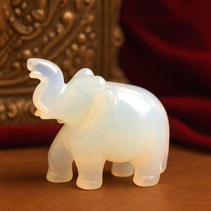 May include: A translucent, off-white elephant figurine. The sculpture has a smooth, polished surface and is carved with detailed features, including the trunk, ears, and legs. The elephant is standing on a brown surface, with a blurred background.