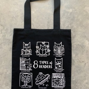 May include: Black tote bag featuring white illustrations of cats reading books. The text reads "8 Types of Readers" and lists different types of readers, such as "The Literary snob", "The Book Juggler", and "The Re-reader".