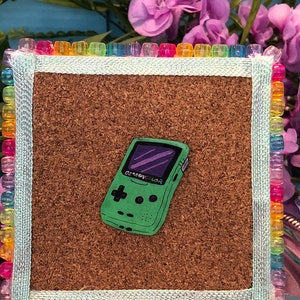 May include: A green Game Boy Color handheld video game console with a purple screen is pinned to a corkboard with a rainbow border. The corkboard is on a small wooden easel.