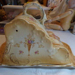 Puede incluir: Un servidor de queso de cerámica vintage con un diseño floral decorativo. El servidor tiene la forma de una cuña de queso y tiene un asa en la parte superior. El servidor está decorado con detalles dorados y tiene un patrón floral en tonos de rosa, amarillo y verde.