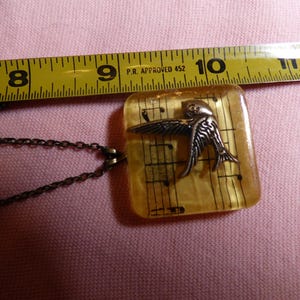 May include: A gold necklace with a square pendant featuring a silver bird flying over a sheet of music. The pendant is about 1 inch wide.