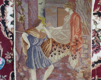 May include: A vintage needlepoint tapestry depicting two figures in a garden setting. The woman on the left is wearing a blue dress and holding a flower. The woman on the right is wearing a red dress and holding a white cloth. The tapestry is framed with a white border.