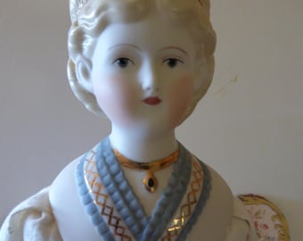May include: A porcelain doll head with blonde hair and blue eyes. The doll wears a gold headband and a blue and gold necklace with a black pendant. The doll is wearing a white dress.