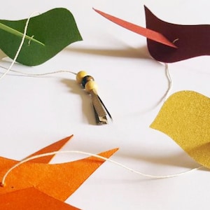 Guirlande artisanale de cinq oiseaux en papier recyclé coloré, montée à la main sur un fil avec perles de bois et clochette décorative. Idéale pour chambre d’enfant, décoration poétique ou fête nature.