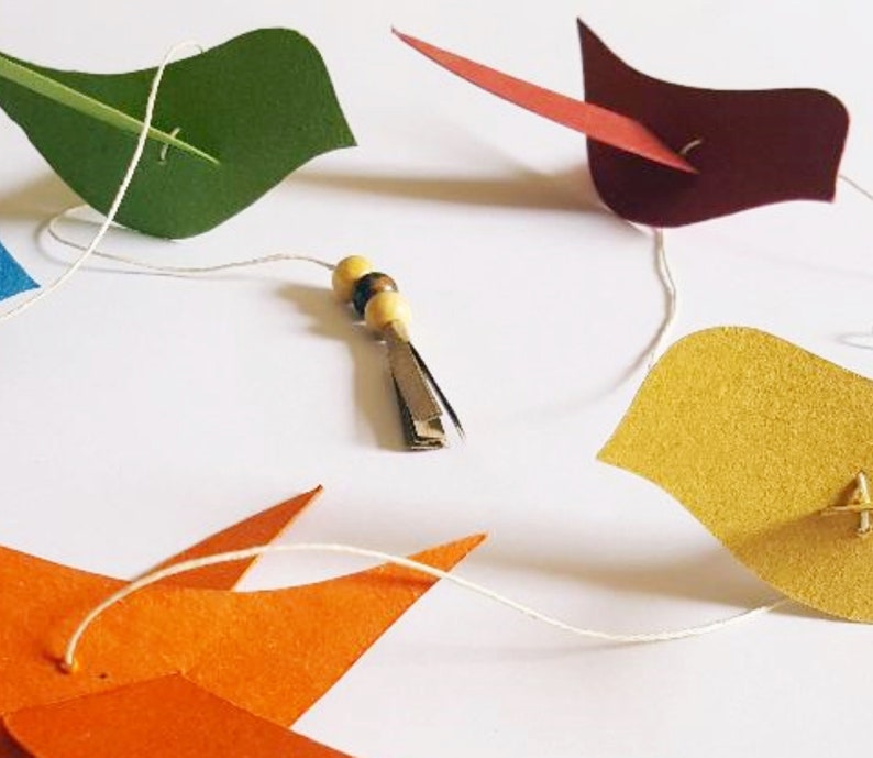 Guirlande artisanale de cinq oiseaux en papier recyclé coloré, montée à la main sur un fil avec perles de bois et clochette décorative. Idéale pour chambre d’enfant, décoration poétique ou fête nature.