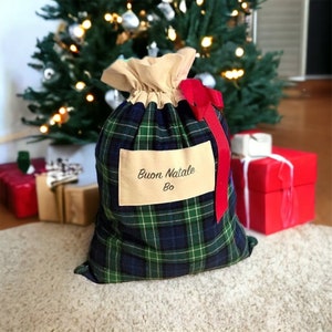 May include: A Christmas gift bag in a green, blue, and white tartan pattern. The bag has a beige drawstring top and a red ribbon. The text "Buon Natale Bo" is on a beige label on the front. A Christmas tree is in the background.