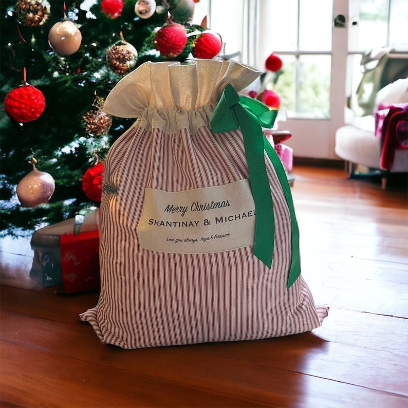 May include: A fabric gift sack with red and white vertical stripes, a cream-coloured top section, and a green ribbon tie. A rectangular patch on the front reads Merry Christmas SHANTINAY & MICHAEL Love you always, Yaya & Pinepaw.