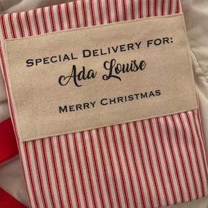 May include: A red and white striped fabric sack featuring a beige rectangular patch with the black printed text SPECIAL DELIVERY FOR: Ada Louise MERRY CHRISTMAS. The sack has a simple rectangular shape with visible fabric texture.