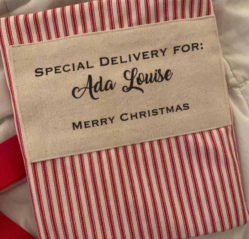 May include: A red and white striped fabric sack featuring a beige rectangular patch with the black printed text SPECIAL DELIVERY FOR: Ada Louise MERRY CHRISTMAS. The sack has a simple rectangular shape with visible fabric texture.