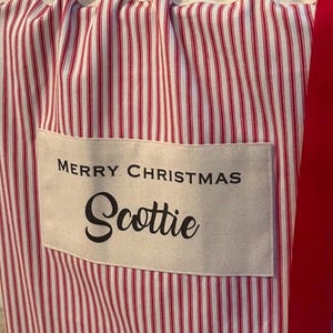 May include: A fabric gift sack featuring a red and white vertical striped pattern. The top is cinched with a red ribbon. A rectangular cream-coloured patch is sewn on the front with the black text MERRY CHRISTMAS and the name Scottie.