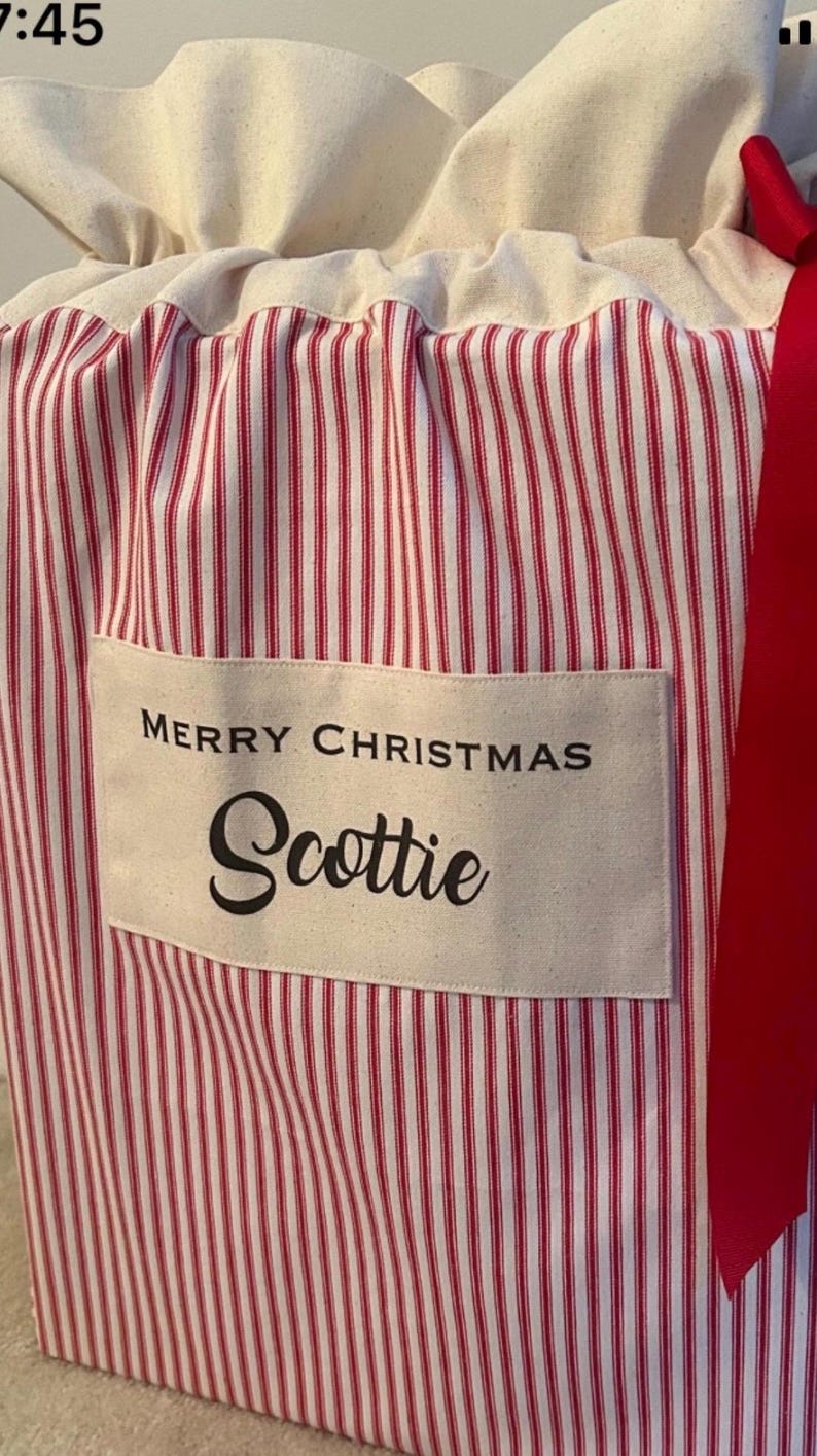 May include: A fabric gift sack featuring a red and white vertical striped pattern. The top is cinched with a red ribbon. A rectangular cream-coloured patch is sewn on the front with the black text MERRY CHRISTMAS and the name Scottie.