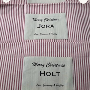 May include: Two fabric items featuring a red and white vertical striped pattern. Each item has a rectangular white patch with the black printed text Merry Christmas, a name, and the phrase Love, Grammy & Pappy, specifically JORA and HOLT.