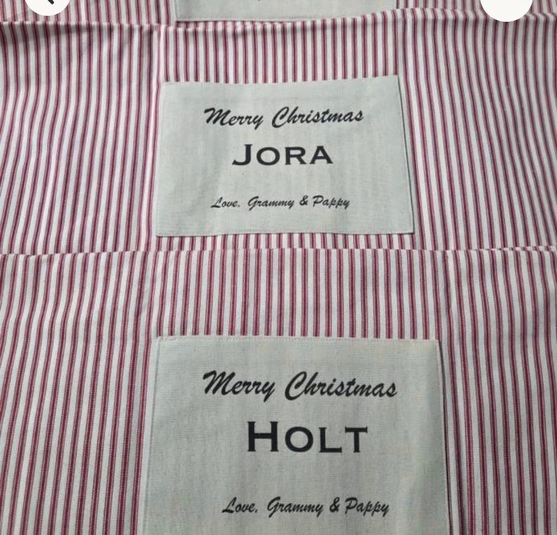 May include: Two fabric items featuring a red and white vertical striped pattern. Each item has a rectangular white patch with the black printed text Merry Christmas, a name, and the phrase Love, Grammy & Pappy, specifically JORA and HOLT.