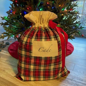 May include: A large Christmas gift bag with a red, black, and cream plaid pattern. The bag has a beige drawstring top and a red ribbon. The name "Cade" is printed on a rectangular label on the front of the bag. A Christmas tree is in the background.