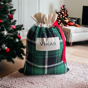 May include: A large Christmas gift bag with a green and black tartan pattern. The bag has a cream drawstring top and a label that reads "VIKAS". A red ribbon is tied to the bag. A Christmas tree is in the background.