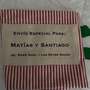 May include: A fabric sack featuring a red and white vertical striped pattern with a rectangular beige patch sewn on the front. The patch displays the black text: ENV&Iacute;O ESPECIAL PARA: MAT&Iacute;AS Y SANTIAGO DE: PAP&Aacute; NOEL Y LOS REYES MAGOS.