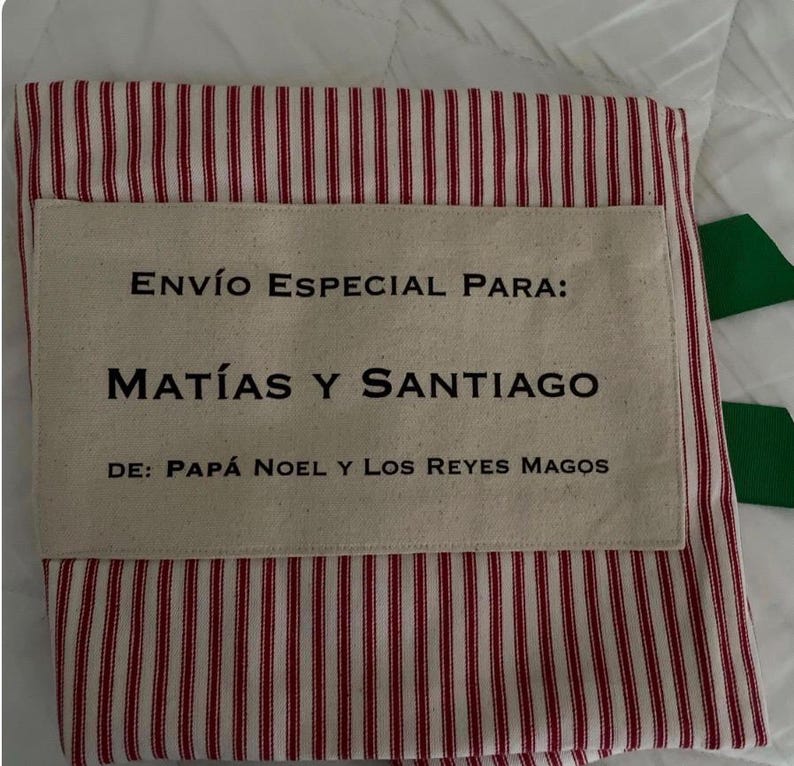 May include: A fabric sack featuring a red and white vertical striped pattern with a rectangular beige patch sewn on the front. The patch displays the black text: ENV&Iacute;O ESPECIAL PARA: MAT&Iacute;AS Y SANTIAGO DE: PAP&Aacute; NOEL Y LOS REYES MAGOS.