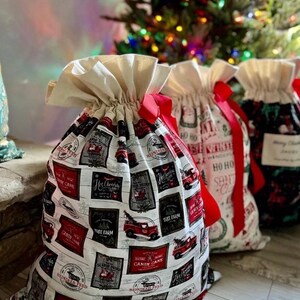 May include: Three fabric drawstring gift bags with ruffled tops and red ribbon closures. The front bag features a white background with a repeating grid pattern of red and black holiday-themed rectangular labels depicting trucks and text.