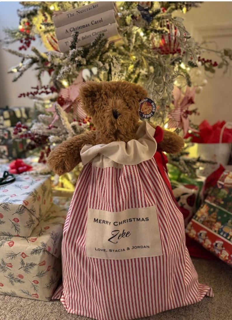 May include: A brown plush teddy bear sits inside a red and white striped fabric gift sack. The sack features a cream-coloured rectangular patch with the text: MERRY CHRISTMAS, Zeke, LOVE, STACIA & JORDAN. A red ribbon is tied to the side.