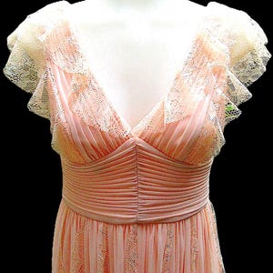 K&ouml;nnte beinhalten: Hellrosa Vintage-Kleid mit V-Ausschnitt und zarten Spitzendetails. Das Kleid hat ger&uuml;schte Fl&uuml;gel&auml;rmel und eine betonte Taille. Der Rock hat ein dezentes Spitzenmuster. Das Kleid wird auf einer Schaufensterpuppe pr&auml;sentiert.