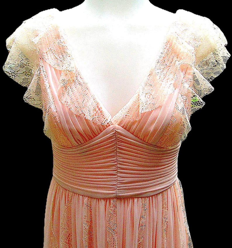 K&ouml;nnte beinhalten: Hellrosa Vintage-Kleid mit V-Ausschnitt und zarten Spitzendetails. Das Kleid hat ger&uuml;schte Fl&uuml;gel&auml;rmel und eine betonte Taille. Der Rock hat ein dezentes Spitzenmuster. Das Kleid wird auf einer Schaufensterpuppe pr&auml;sentiert.