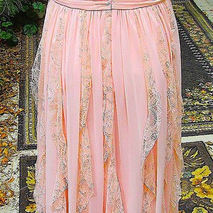 K&ouml;nnte beinhalten: Ein bodenlanges, rosa Vintage-Kleid mit zarten Spitzenbes&auml;tzen. Das Kleid hat einen V-Ausschnitt, eine betonte Taille und flie&szlig;ende Rockbahnen mit Spitzenakzenten. Das Kleid ist aus einem weichen, flie&szlig;enden Stoff gefertigt.